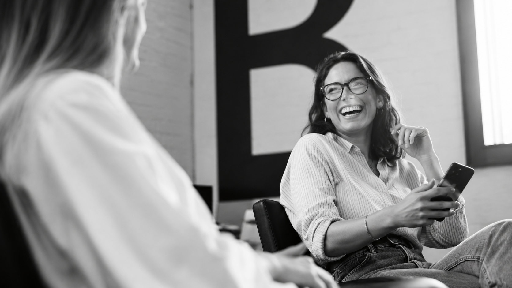 Black-and-white image of two professionals laughing during a casual office conversation.