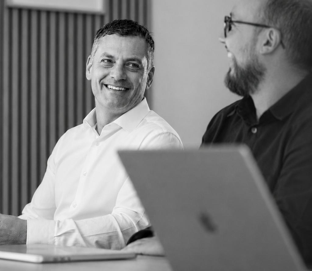 Two colleagues smiling during a meeting, seated with laptops as they discuss ideas in a modern office.