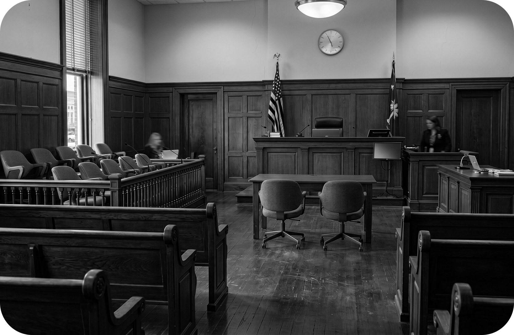Black and white image of the view of a state courtroom featuring pews, chairs, a desk, flags, and the stand. Figures in motion move about the room.