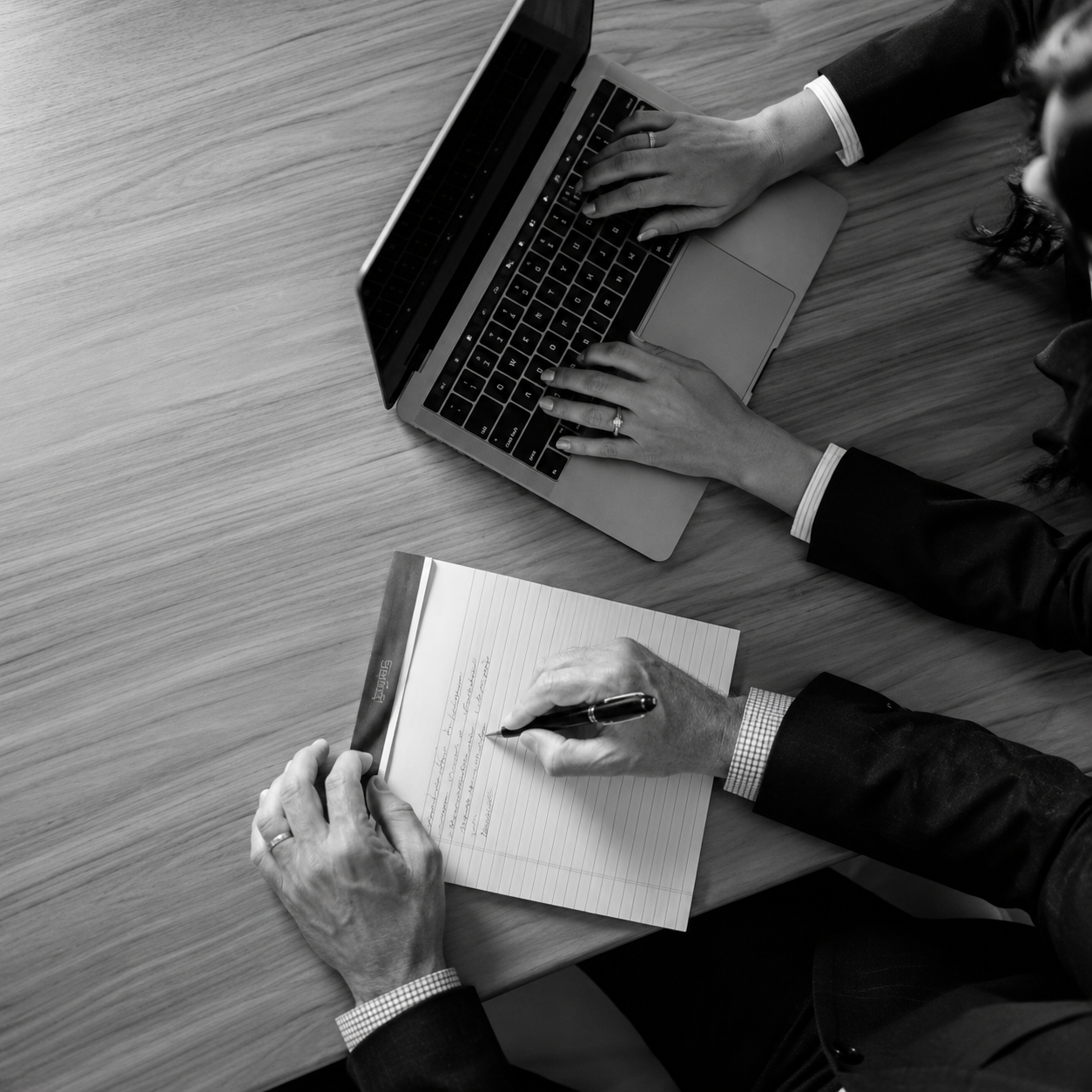 Overhead view of two professionals collaborating, with one typing on a laptop while the other takes notes during a business meeting.