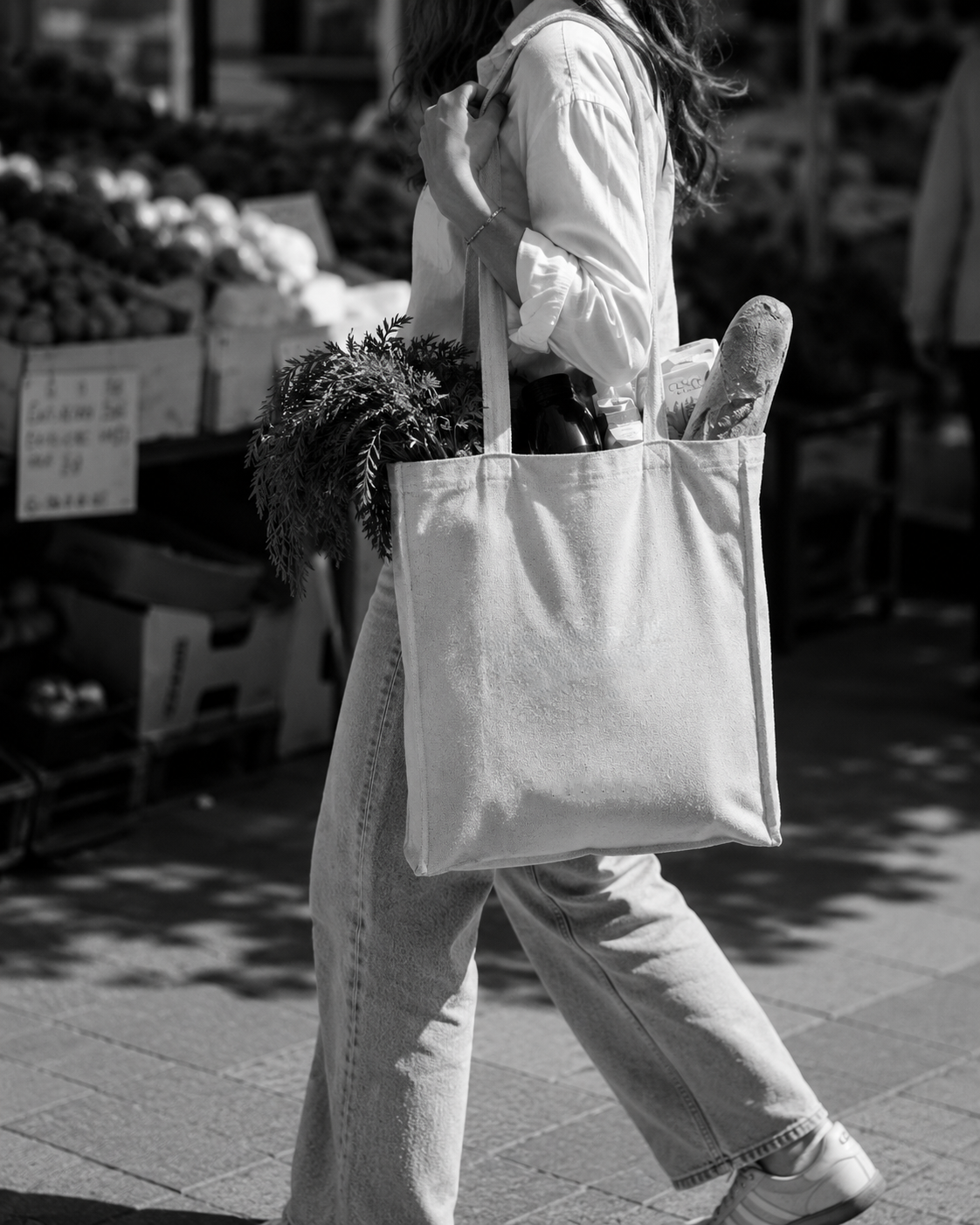 Black-and-white image of a shopper carrying a tote bag filled with fresh groceries and consumer goods.