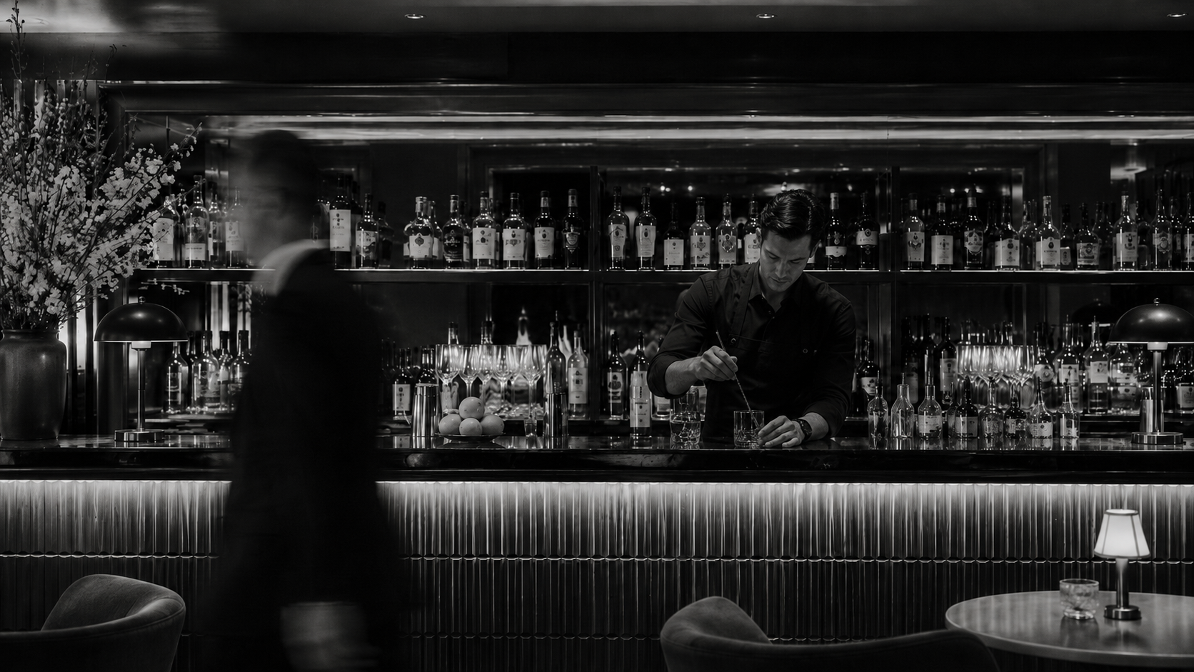 Black-and-white image of a bartender preparing drinks behind an upscale hotel bar.