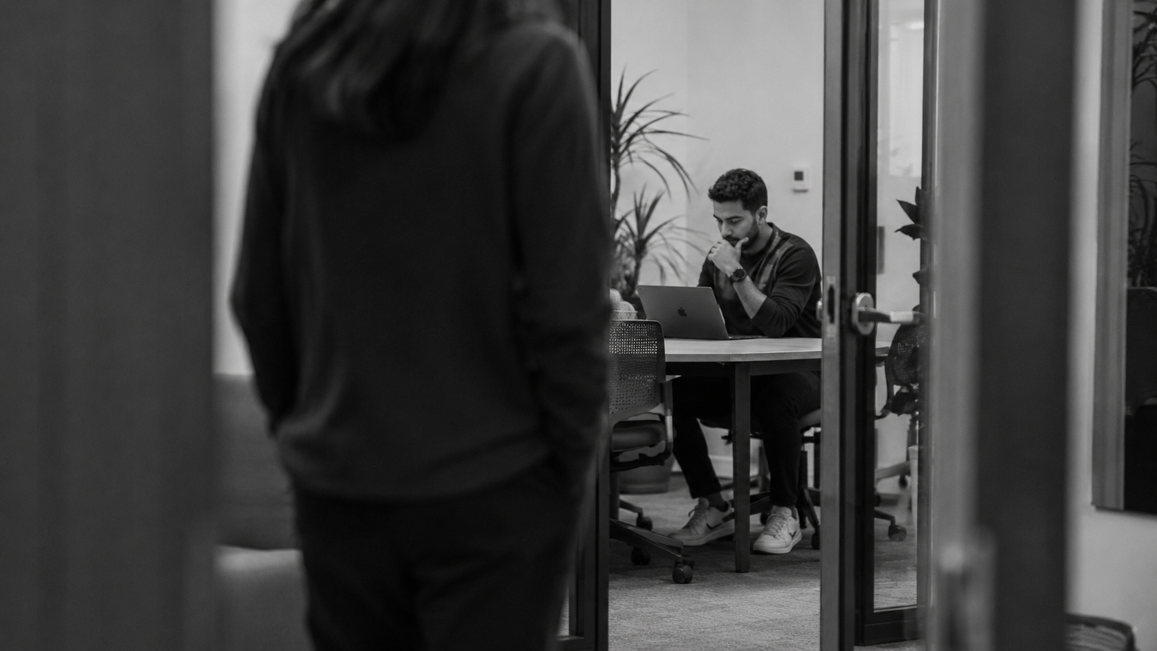 Man focused on laptop in busy, modern coworking space. A woman passes in front of the room he is working in.