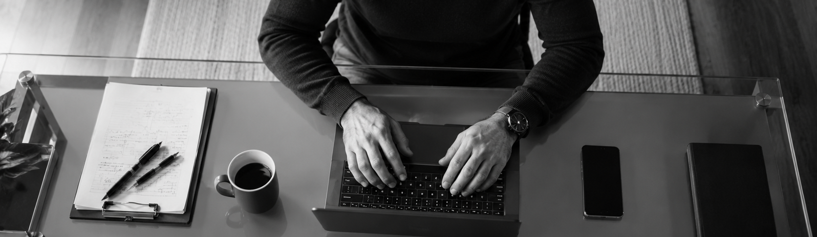 Black-and-white image of a business owner working on a laptop at a desk with notes, coffee, and a phone nearby.